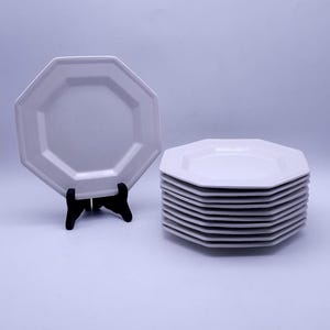 Puede incluir: Un conjunto de platos blancos octogonales con borde de cuentas, junto a un plato individual sobre un soporte negro. Los platos son de color blanco brillante y parecen ser de cerámica o porcelana. Diseño sencillo y elegante.