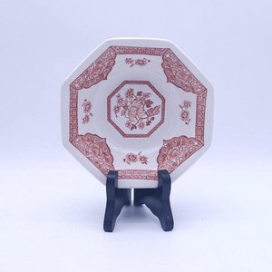 Op de afbeelding: Een achthoekig keramieken schoteltje met een witte basis en een roodbruin bloemmotief. Het ontwerp bevat een centraal boeket binnen een achthoekige rand en decoratieve bloemmotieven langs de rand, gepresenteerd op een zwarte kunststof standaard.