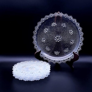 Op de afbeelding: Transparante glazen serveerschaal met een bubbelachtig ontwerp en een bijpassende stapel onderzetters. De schaal wordt gepresenteerd op een donkere houten standaard tegen een zwarte achtergrond. De schaal en onderzetters hebben een decoratieve, geschulpte rand.