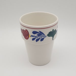 Op de afbeelding: Een witte keramische beker met een rood, blauw en groen bloemmotief rond de rand.