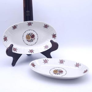 Op de afbeelding: Twee witte ovale keramische schalen met een geschulpte rand en een centraal bloemmotief van gele en bordeauxrode bloemen omringd door een decoratieve rand. Kleine bordeauxrode bloemtakjes zijn gelijkmatig verdeeld over de rand.
