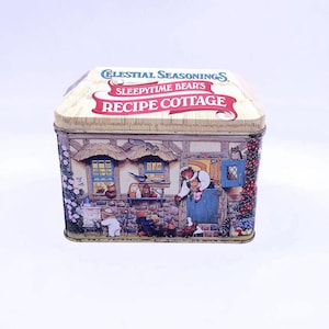 Puede incluir: Una caja de recetas rectangular de metal con una escena de cabaña con osos. La caja tiene una tapa de color crema con el texto "Celestial Seasonings Sleepytime Bear's Recipe Cottage" en rojo y azul. Los lados muestran una ilustración detallada de una cabaña.