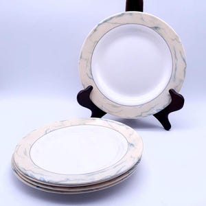 May include: Set of four white ceramic dinner plates with a light blue and beige marble design. The plates have a gold rim.
