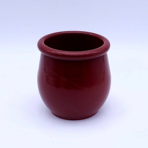 Puede incluir: Una maceta de cerámica de color rojo oscuro con forma redondeada y un borde ligeramente acampanado.