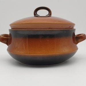May include: A ceramic casserole dish with a lid. The dish has a brown and black gradient colour scheme. It features two side handles and a loop handle on the lid. The dish is designed for cooking and serving.