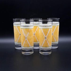 Op de afbeelding: Een set van vijf heldere glazen met een geel ruitpatroon, witte kruisende lijnen en blauwe en groene geometrische randen aan de boven- en onderkant. De hoge, cilindervormige drinkglazen hebben een gladde en transparante afwerking.
