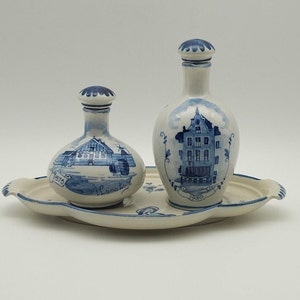 Op de afbeelding: Een blauw en wit keramisch dienblad met twee kleine blauwe en witte keramische flessen. De flessen hebben afbeeldingen van gebouwen en de data 1970 en 1950.