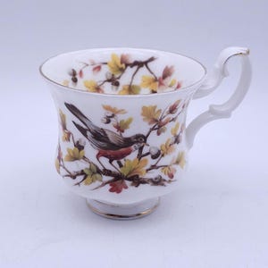 Puede incluir: Una taza de té de porcelana blanca con una ilustración detallada de un pájaro posado en una rama con hojas de otoño amarillas y marrones y pequeñas bayas blancas. La taza tiene un borde festoneado, un asa curva decorativa y base dorada.