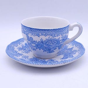 Op de afbeelding: Een witte theekop en bijpassende schotel van keramiek met een gedetailleerd blauw bloemenpatroon. Het ontwerp bevat complexe bloesems en bladeren op de buitenkant van de kop en de schotel, afgewerkt met een decoratieve blauwe kantachtige rand langs de bovenkant van de kop.
