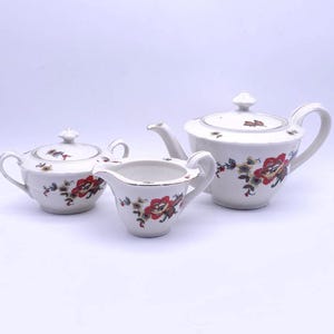 May include: A white porcelain tea set featuring a teapot, sugar bowl, and creamer. Each piece is adorned with a floral pattern in red, yellow, and green. The teapot has a lid and a curved handle, while the creamer has a spout.