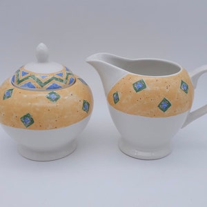May include: A white ceramic sugar bowl and creamer set with a yellow, green, and blue geometric pattern. The lid of the sugar bowl has a blue and green chevron pattern.