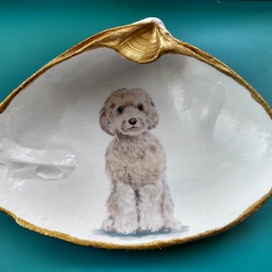 May include: A white seashell with a gold rim, painted with a light brown dog sitting inside. The dog has a white chest and paws.