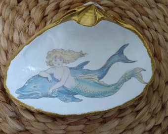 May include: A white ceramic seashell dish with a gold rim, featuring a painted image of a mermaid riding a dolphin. The mermaid has long blonde hair and a blue tail.