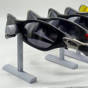 May include: A display of several pairs of sunglasses with black frames and dark lenses. The sunglasses are on gray stands. Some of the sunglasses have colored accents, including red, orange, and silver.