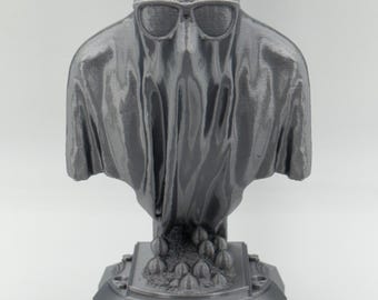 May include: A gray 3D-printed ghost figurine wearing sunglasses. The ghost has a draped appearance and sits atop a detailed base with a rectangular front. The base features a cluster of small, round shapes.