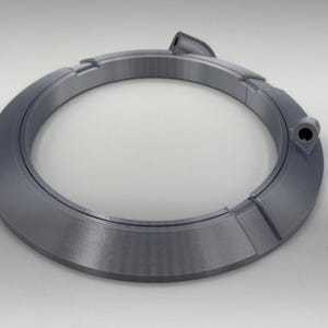 May include: A gray, circular object with a segmented design. The object has a raised section and a small hole. The item is made of a solid material and sits on a white surface. The object appears to be a component or part.