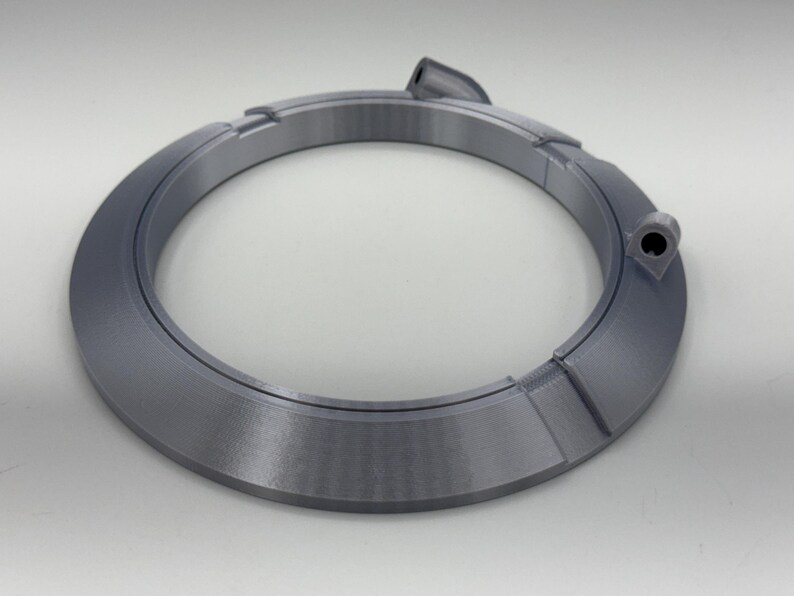 May include: A gray, circular object with a segmented design. The object has a raised section and a small hole. The item is made of a solid material and sits on a white surface. The object appears to be a component or part.