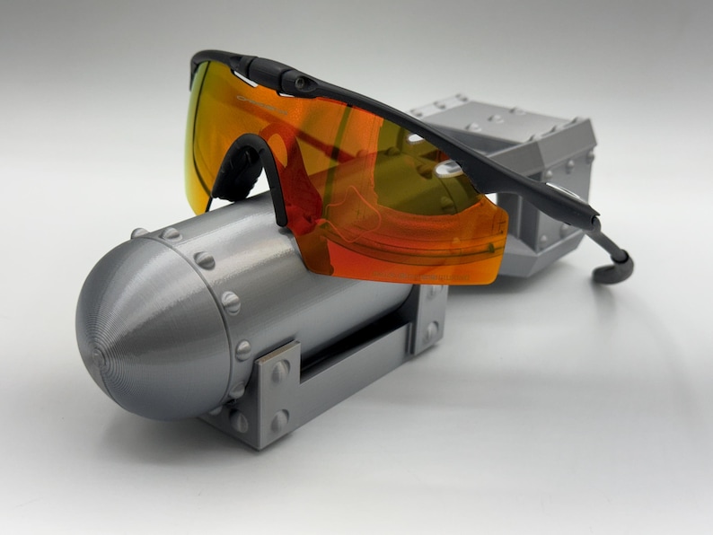 May include: Black sports sunglasses with orange-tinted lenses rest on a gray, cylindrical object with rivets and a rectangular base. The sunglasses have a sleek, modern design, with a black frame and curved temples.