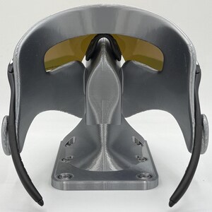May include: A gray and black sports mask with yellow tinted lenses. The mask has a curved design with a nose bridge and black ear pieces. The mask is mounted on a gray stand with a rectangular base.