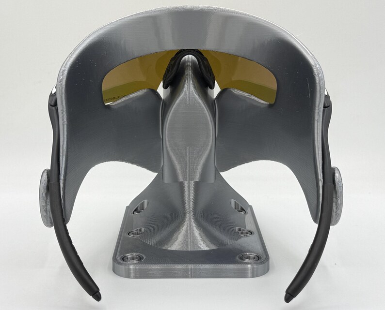 May include: A gray and black sports mask with yellow tinted lenses. The mask has a curved design with a nose bridge and black ear pieces. The mask is mounted on a gray stand with a rectangular base.