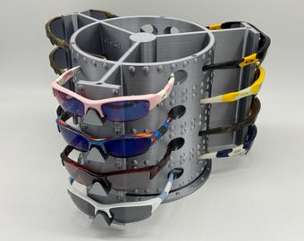 May include: A silver, cylindrical, rotating display stand featuring multiple vertical slots designed to hold various pairs of sunglasses. The stand has a textured, rivet-like pattern along its exterior and holds several pairs of sports-style eyewear.