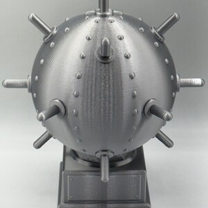 May include: A grey, spherical object with protruding cylindrical elements and small, rounded studs. The object is mounted on a rectangular base. The design suggests a mechanical or industrial theme.