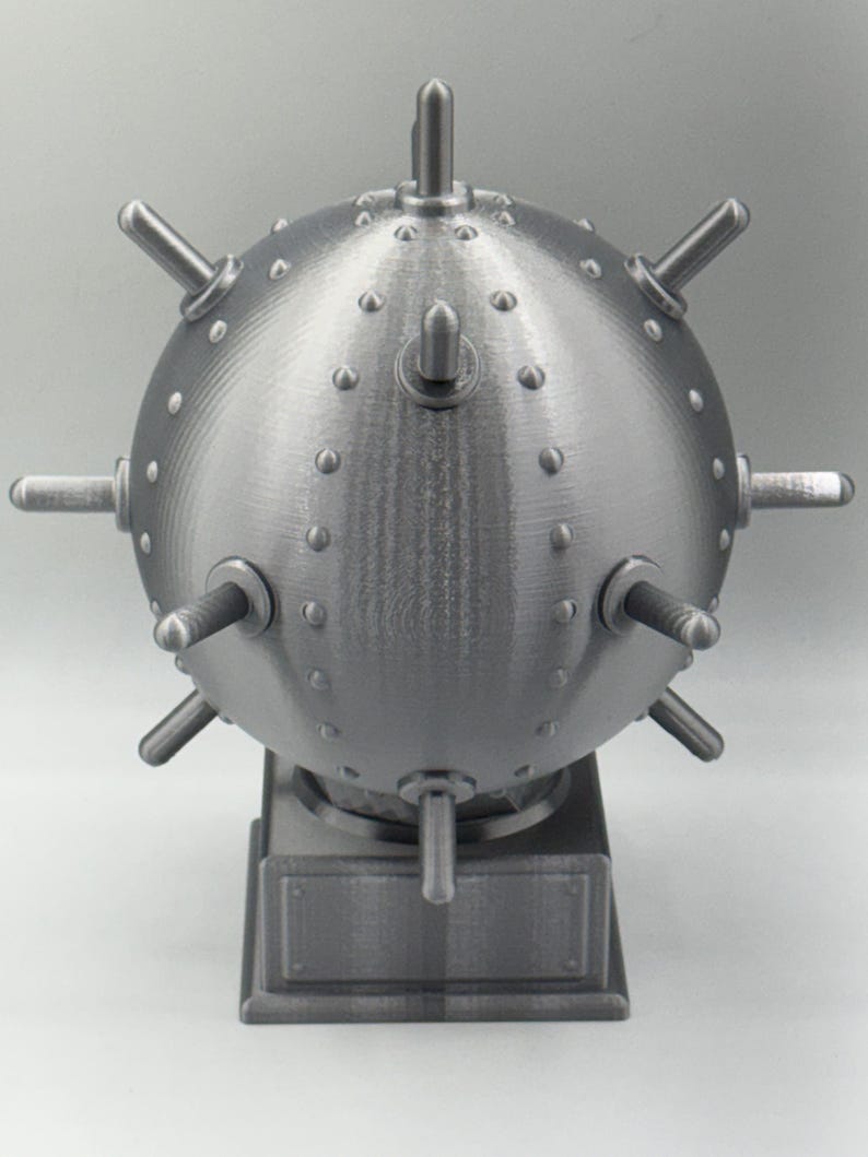 May include: A grey, spherical object with protruding cylindrical elements and small, rounded studs. The object is mounted on a rectangular base. The design suggests a mechanical or industrial theme.
