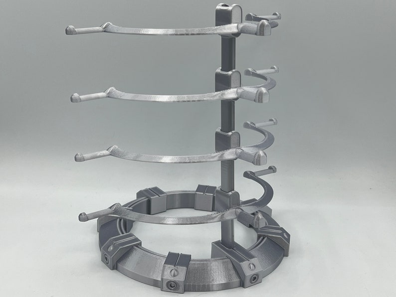 May include: A silver metal stand with four tiers for displaying items. Each tier has a curved arm with a rounded end. The stand has a circular base with a textured surface.