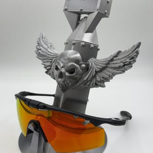 May include: A gray 3D-printed sunglasses stand featuring a skull with wings, a bomb-shaped body, and a rectangular top. The stand holds a pair of black sunglasses with orange lenses.
