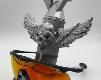 May include: A gray 3D-printed sunglasses stand featuring a skull with wings, a bomb-shaped body, and a rectangular top. The stand holds a pair of black sunglasses with orange lenses.