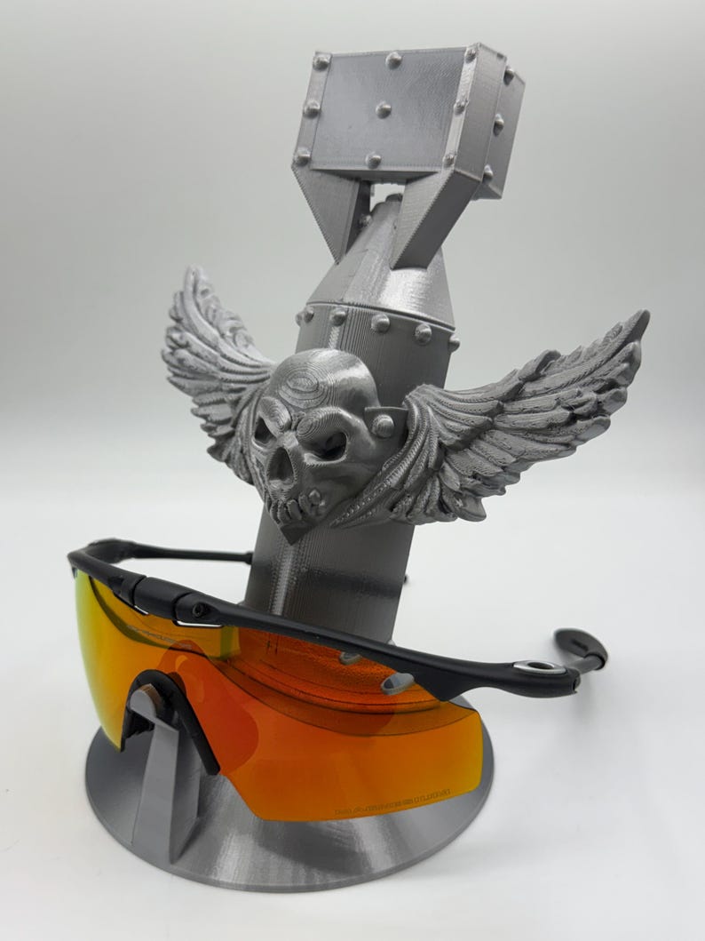 May include: A gray 3D-printed sunglasses stand featuring a skull with wings, a bomb-shaped body, and a rectangular top. The stand holds a pair of black sunglasses with orange lenses.