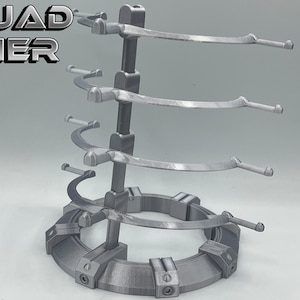 May include: A silver metal stand with four tiers for displaying items. Each tier has a curved arm for holding objects. The stand has a circular base with a textured surface. The stand is labeled "QUAD TIER".