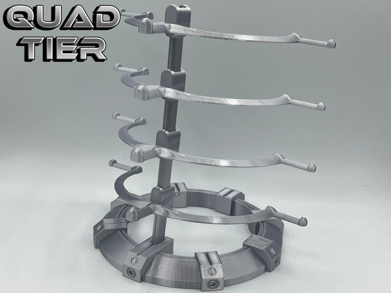 May include: A silver metal stand with four tiers for displaying items. Each tier has a curved arm for holding objects. The stand has a circular base with a textured surface. The stand is labeled "QUAD TIER".