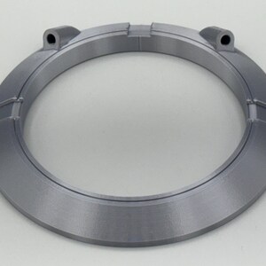 May include: A circular, gray, 3D-printed object with a ring shape. The object has a series of rectangular segments around the outer edge and two small cylindrical protrusions. The object is made of a matte, light gray material.