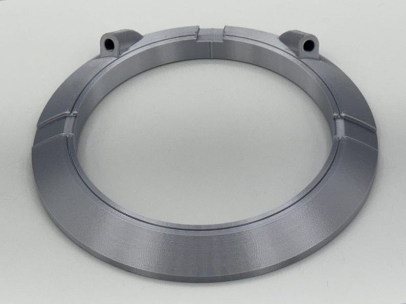 May include: A circular, gray, 3D-printed object with a ring shape. The object has a series of rectangular segments around the outer edge and two small cylindrical protrusions. The object is made of a matte, light gray material.