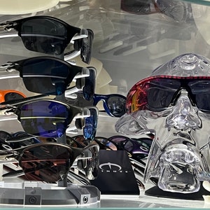 May include: A display of sunglasses with various colored lenses and frames. The sunglasses are arranged on a shelf with a clear plastic head form wearing a pair of sunglasses with a red and black frame.