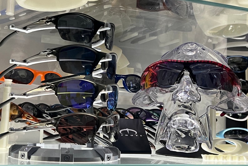 May include: A display of sunglasses with various colored lenses and frames. The sunglasses are arranged on a shelf with a clear plastic head form wearing a pair of sunglasses with a red and black frame.