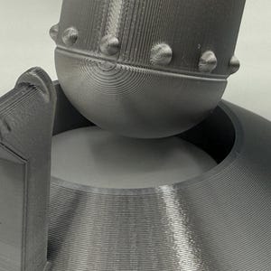 May include: Close-up of a gray, 3D-printed object with a rounded top and a circular base. The object has a textured surface with visible layer lines. The top features small, rounded protrusions.