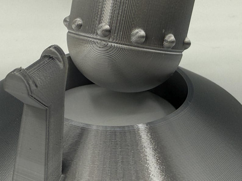 May include: Close-up of a gray, 3D-printed object with a rounded top and a circular base. The object has a textured surface with visible layer lines. The top features small, rounded protrusions.