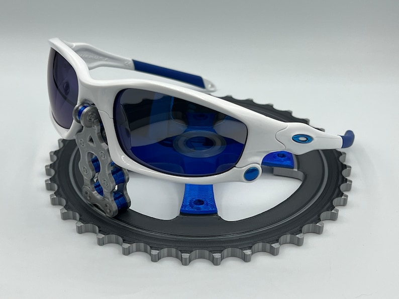 Pu&ograve; includere: Occhiali da sole bianchi con lenti blu e dettagli blu che riposano su un piatto catena per bicicletta nero.