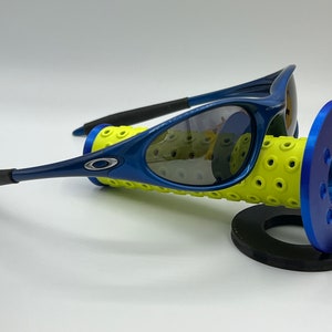 May include: Blue and black Oakley sunglasses with gray lenses. The sunglasses are resting on a yellow and blue display stand.