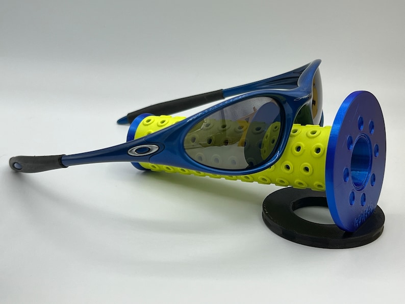 May include: Blue and black Oakley sunglasses with gray lenses. The sunglasses are resting on a yellow and blue display stand.