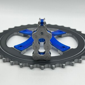 Pu&ograve; includere: Un modello stampato in 3D di una catena per bicicletta, con uno schema di colori blu e grigio, &egrave; mostrato su un pignone nero.
