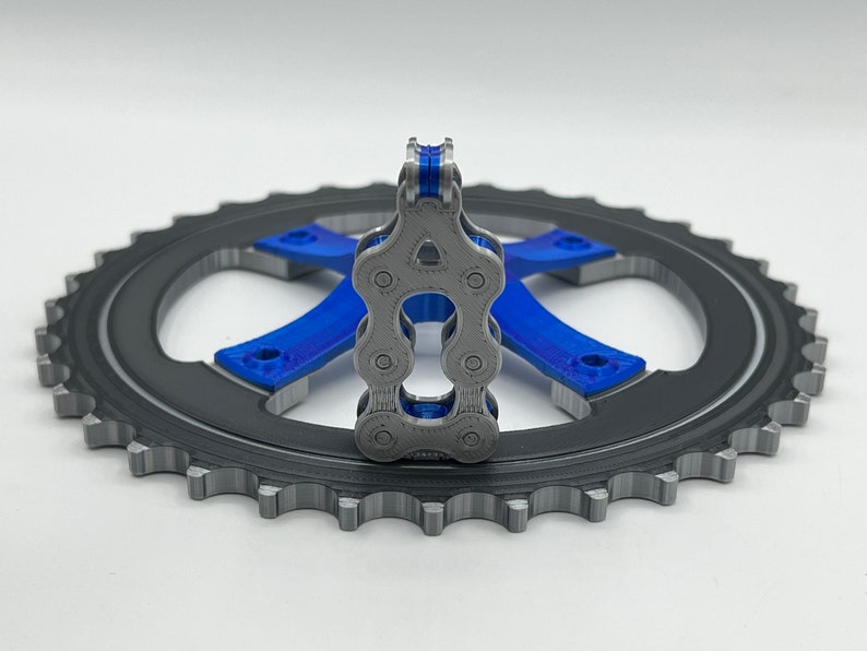 Pu&ograve; includere: Un modello stampato in 3D di una catena per bicicletta, con uno schema di colori blu e grigio, &egrave; mostrato su un pignone nero.