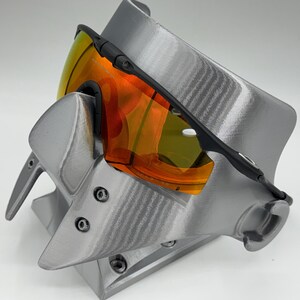 May include: Black-framed sports sunglasses with orange and yellow lenses rest on a gray display stand. The stand has a textured surface and a unique, angular design. The sunglasses are angled, showcasing the vibrant lens colors.