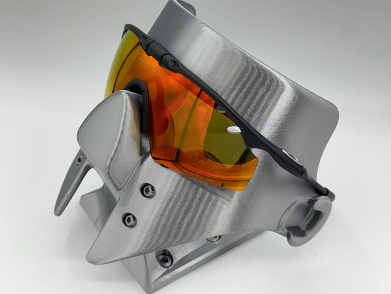May include: Black-framed sports sunglasses with orange and yellow lenses rest on a gray display stand. The stand has a textured surface and a unique, angular design. The sunglasses are angled, showcasing the vibrant lens colors.