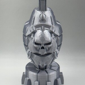 May include: A gray, 3D-printed sculpture of a stylized skull-faced grenade. The sculpture has a textured surface and a cylindrical base with circular details. The top features a rectangular handle.