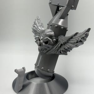 May include: A gray 3D-printed sculpture of a bomb with a skull and wings. The bomb is mounted on a circular base with a support structure. The skull has detailed features, and the wings are spread out. The sculpture is made of a solid, matte material.