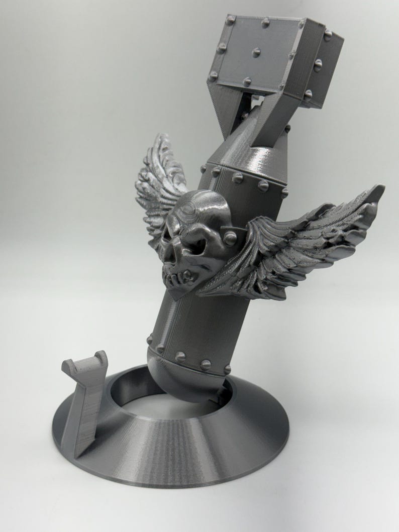 May include: A gray 3D-printed sculpture of a bomb with a skull and wings. The bomb is mounted on a circular base with a support structure. The skull has detailed features, and the wings are spread out. The sculpture is made of a solid, matte material.