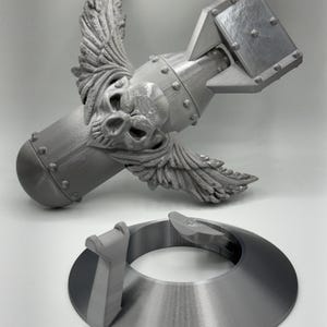 May include: A gray, 3D-printed model of a bomb with a skull and wings, and a separate circular base. The bomb has a detailed skull design and wing accents. The base is a ring with a raised section.